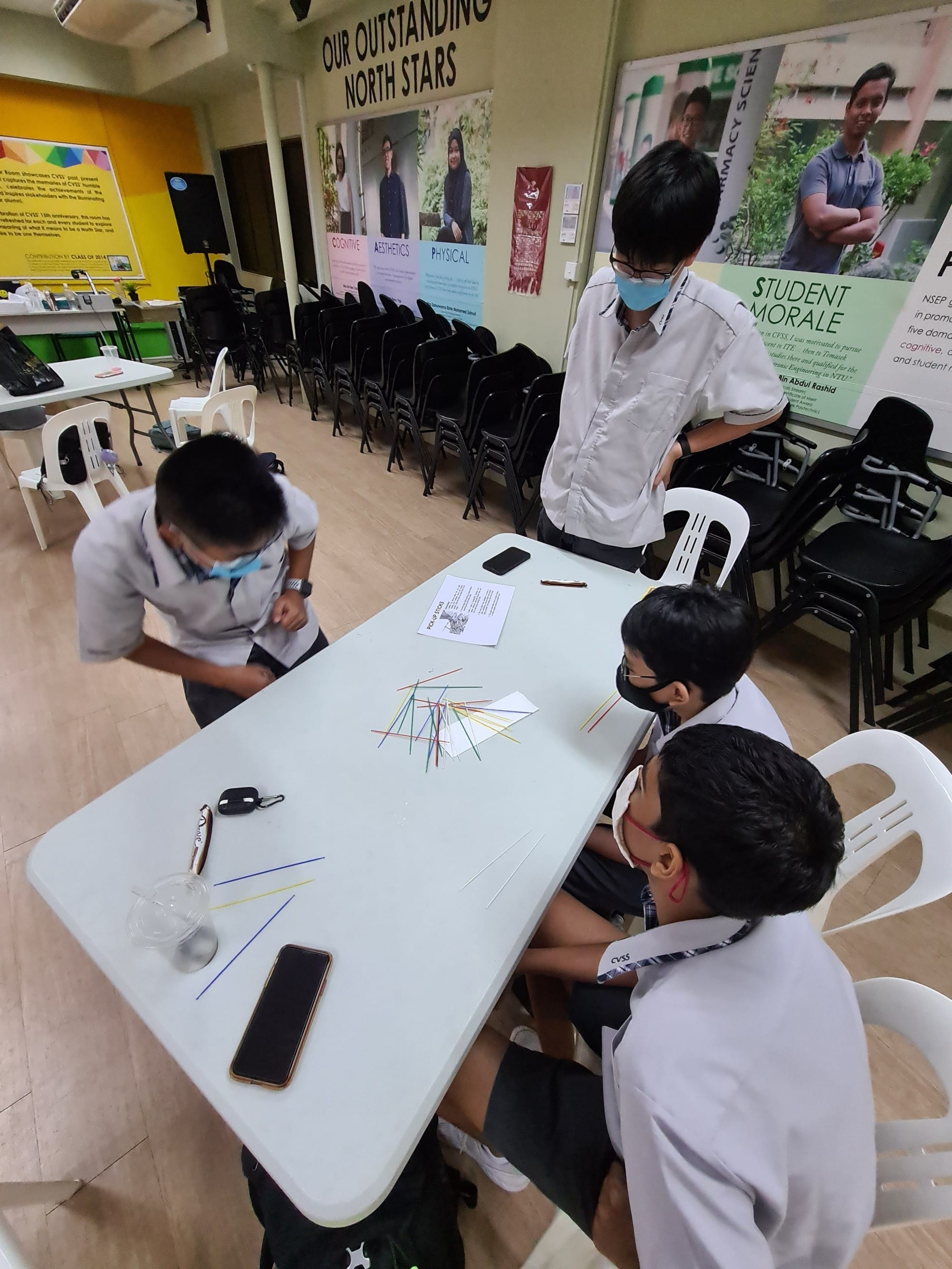 Four masked school students gather around a white table playing pick-up sticks in a decorated school hall.